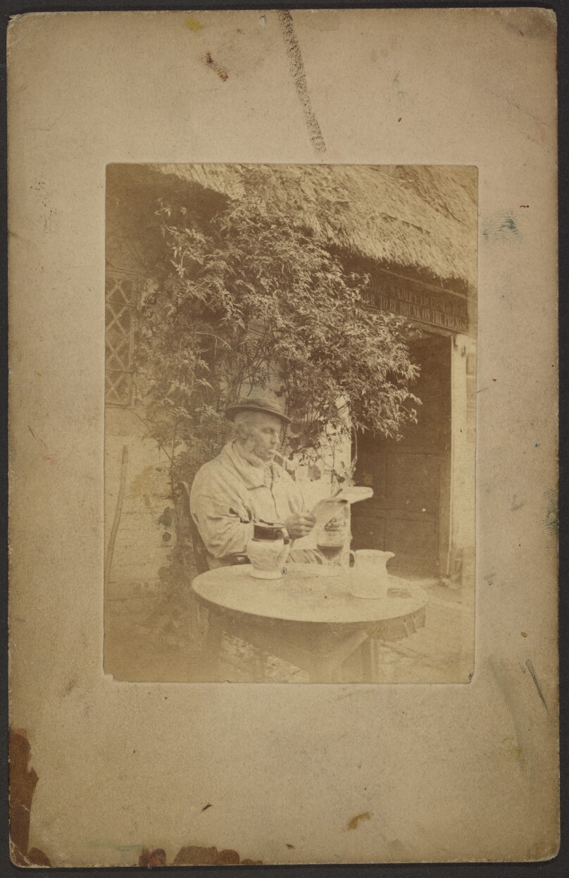 [Man smoking pipe seated at round table in front of tavern]