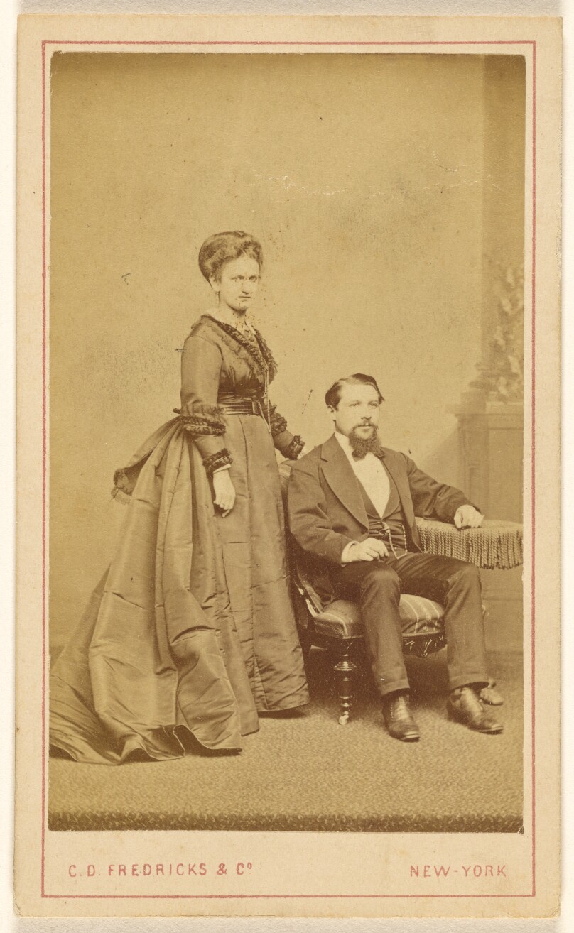 [Unidentified couple: woman standing, man with Vandyke beard seated]