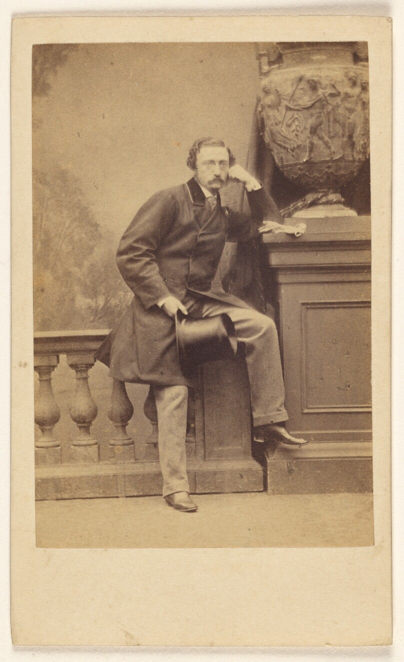 [Man with moustache, holding a top hat, standing, posed on railing in studio]
