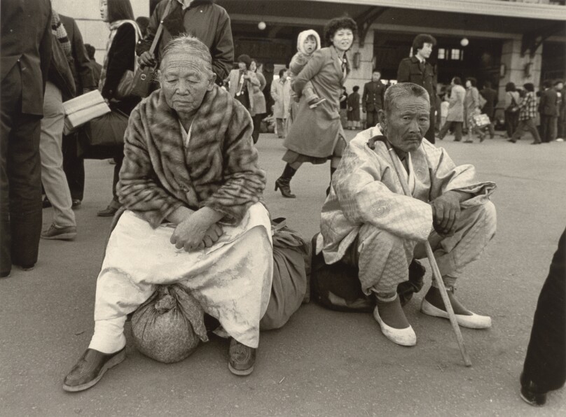Older couple in traditional dress seated on bundles waiting in Seoul Station