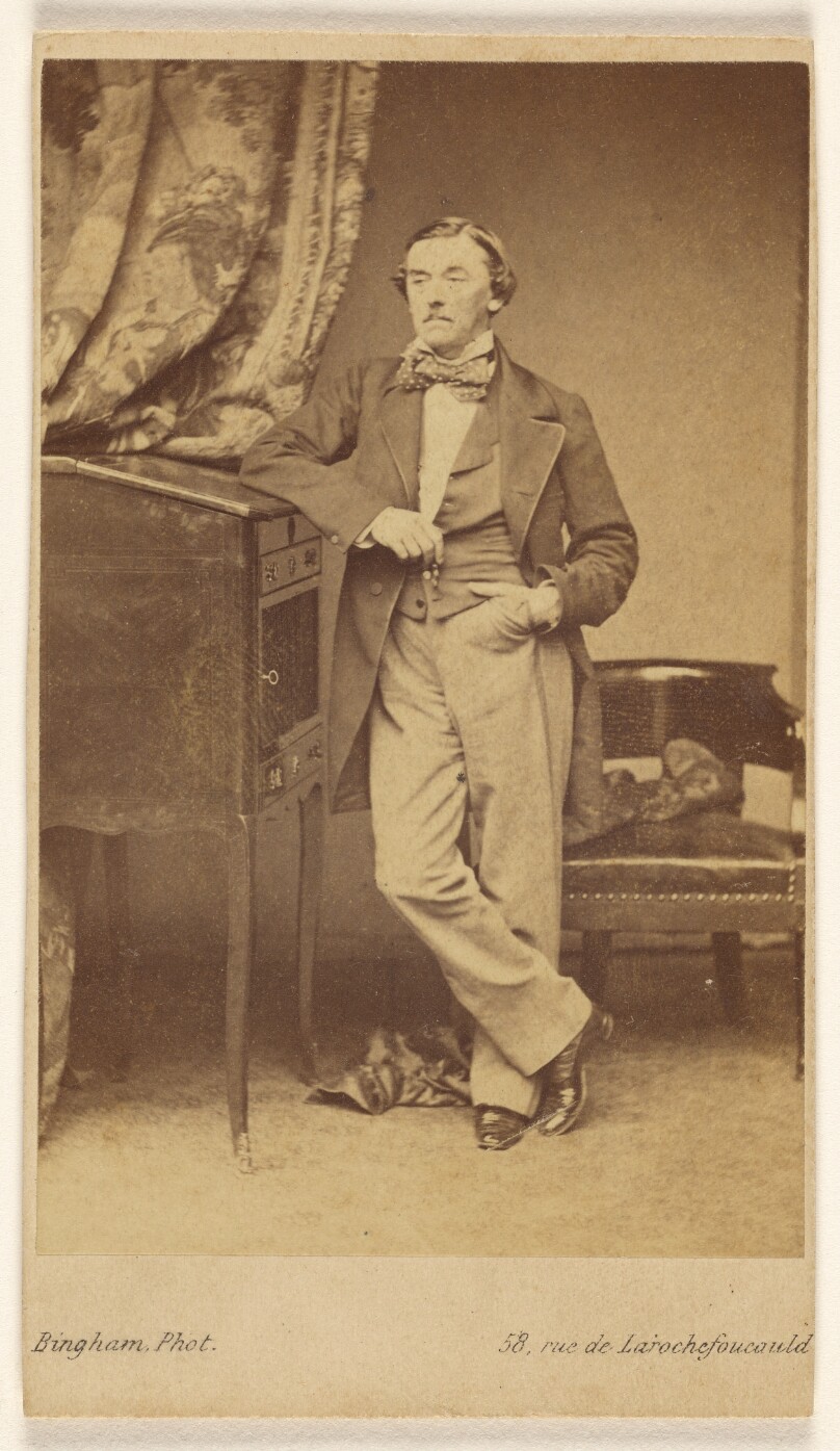 [Unidentified man with moustache wearing a bow tie, standing, with one hand in pocket]