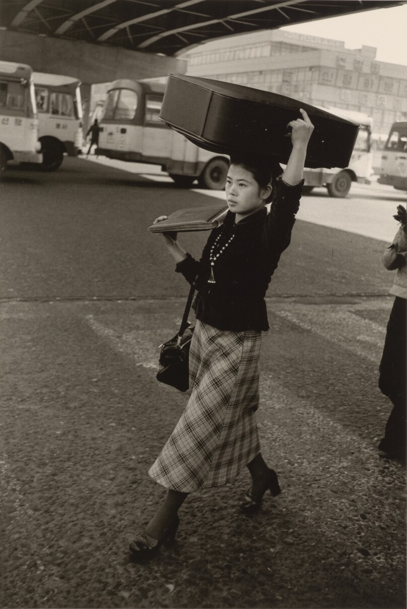 [Young Woman in Plaid Skirt Carrying a Suitcase on Her Head]