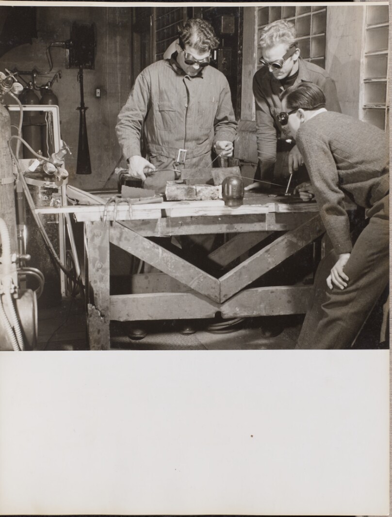 [Three men wearing goggles weld a piece of metal on a table]