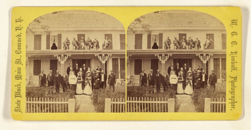 [Large group of people in front of house, some seated on roof]