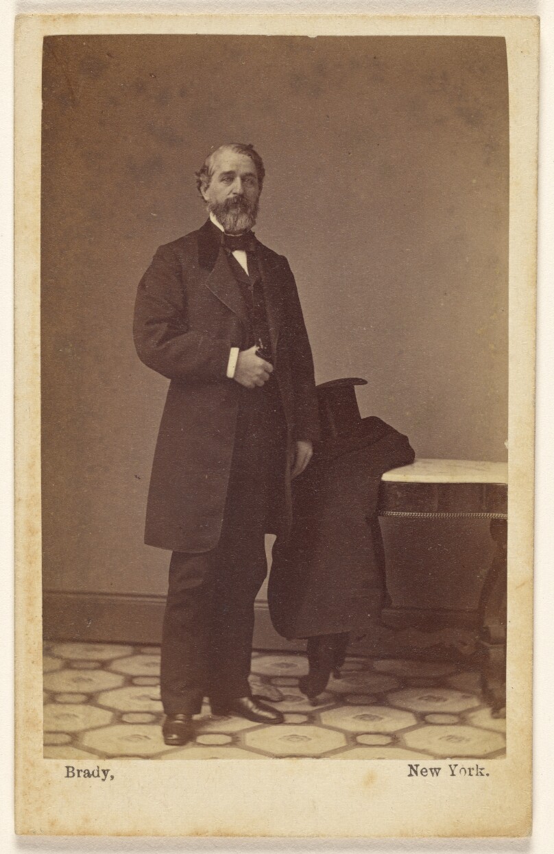 [Unidentified bearded man standing, a top hat and coat on nearby table]