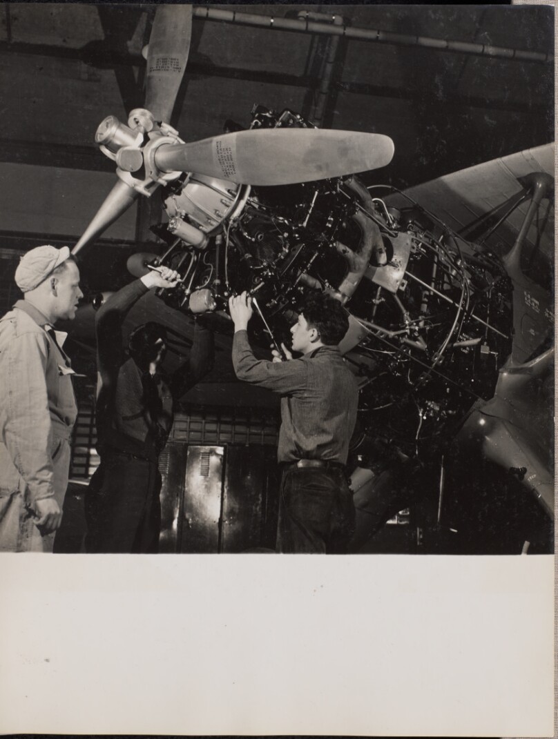 [Three men work on an airplane engine]