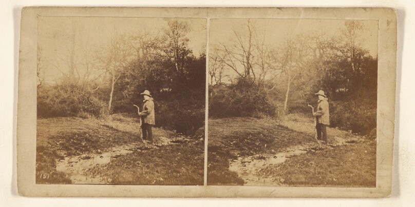 [Man in hat walking down path holding a crooked stick]