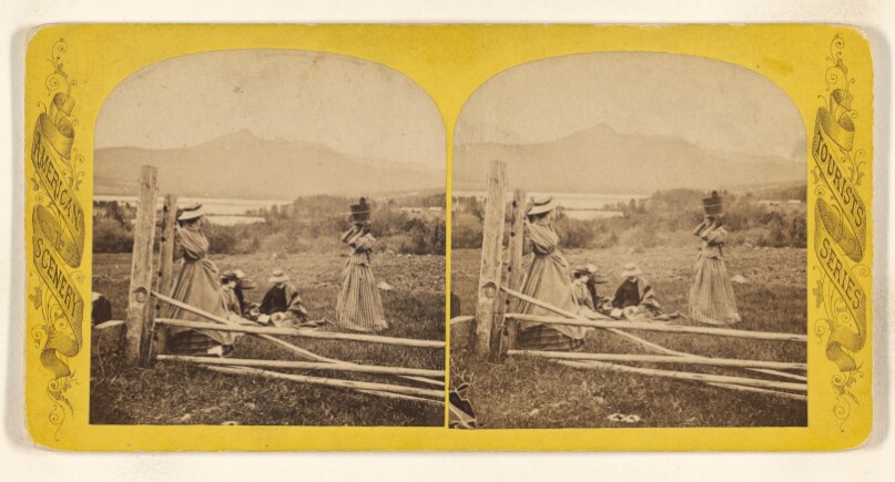[Picnic near lakeside, one woman standing at a fence post, three others seated on ground, one black woman with bucket on head]