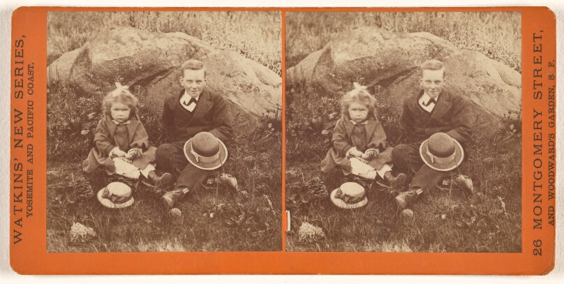 [Unidentified girl and boy seated on grass in front of large rock]