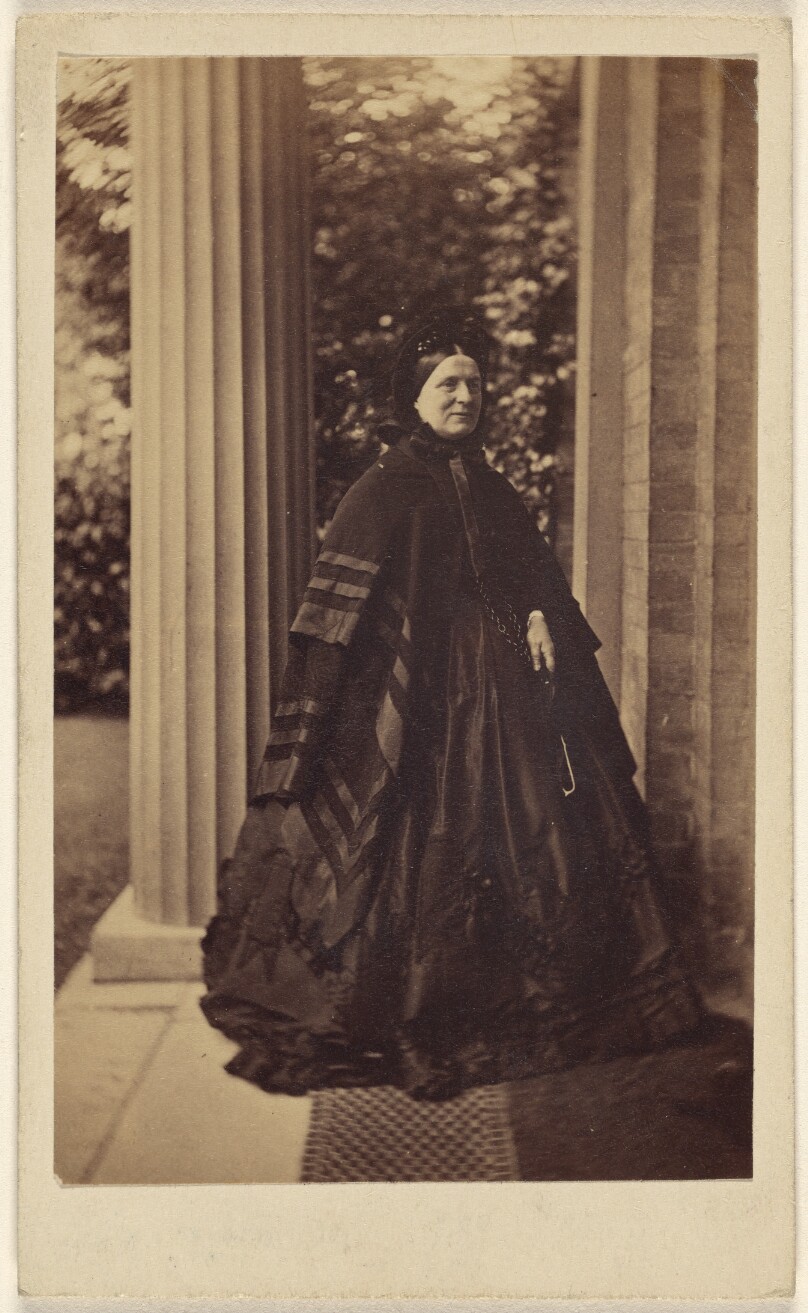 [Unidentified elderly woman standing outside near column at house entrance]