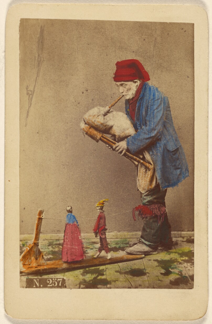 [Unidentified man in cap playing a bagpipe-type instrument with two marionettes]