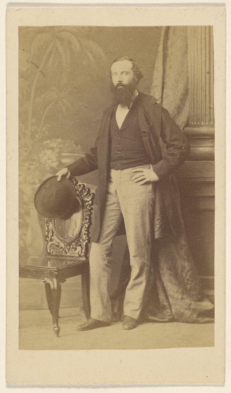 [Unidentified bearded man standing with one hand on hip, the other with hat in hand on chair back]
