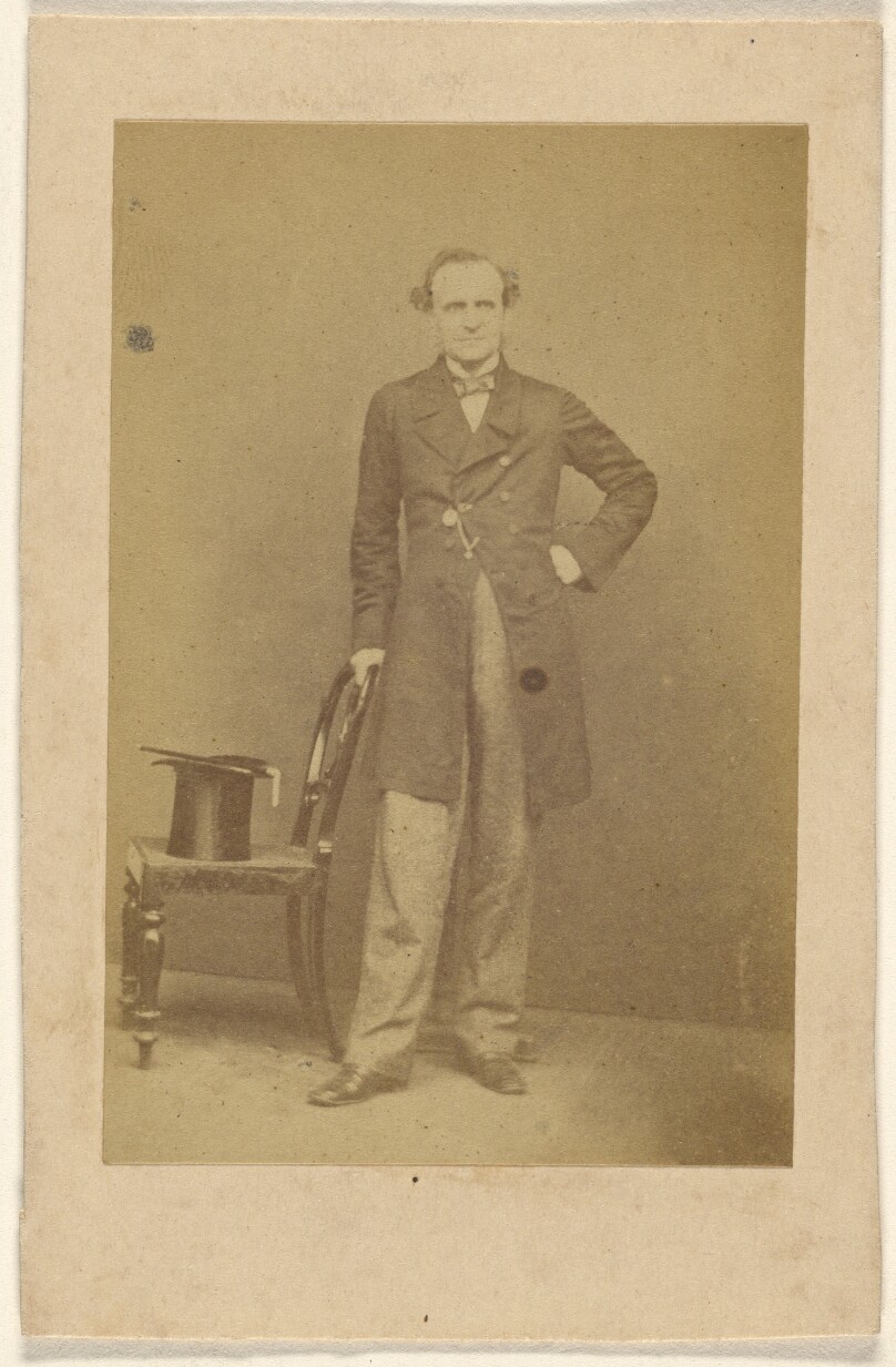 [Unidentified man standing, holding chair back with top hat in seat]