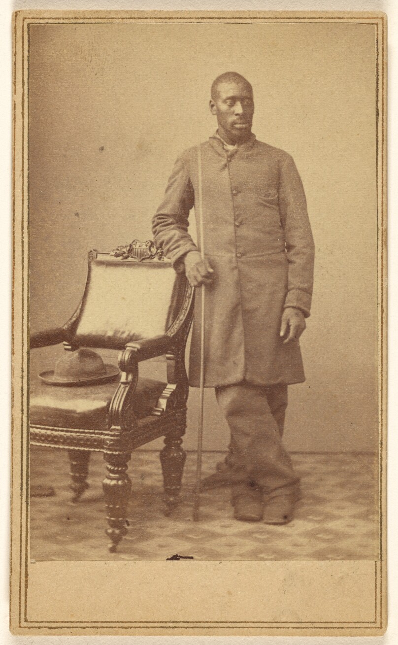 [Black soldier standing near chair with hat in seat]