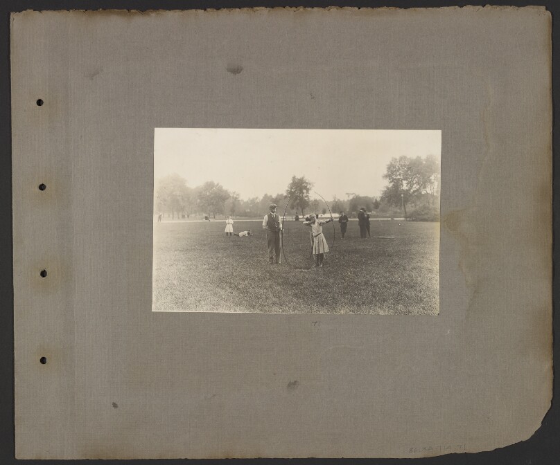 [Archery field, old man and young girl shooting in foreground]