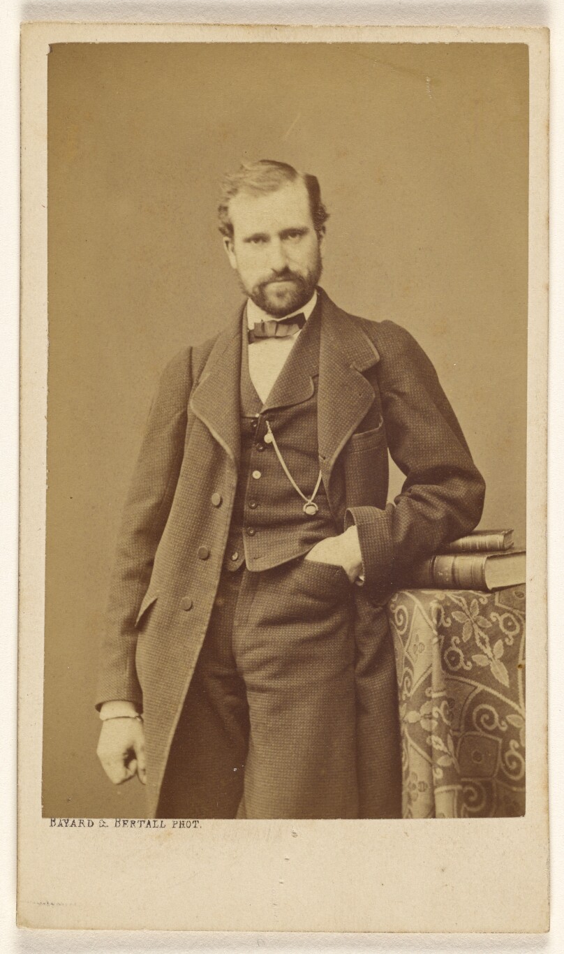 [Unidentified well-dressed bearded man standing, elbow on books on a table]