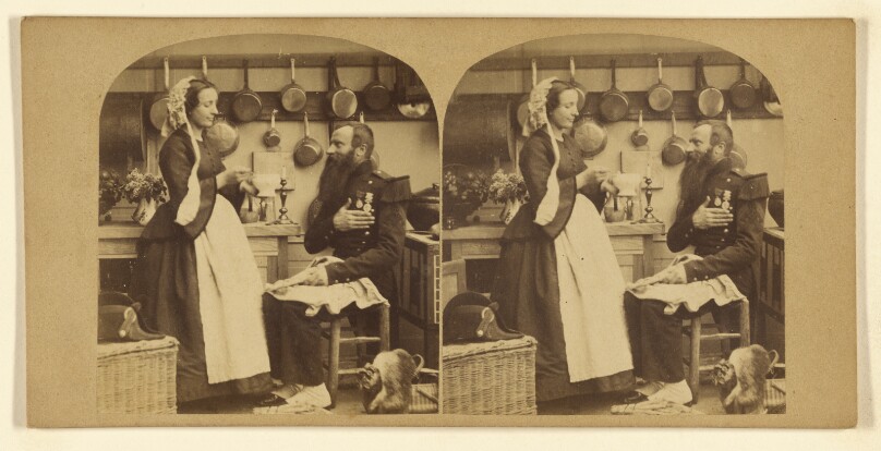 [Bearded man in military garb seated being fed soup by a woman in a kitchen]