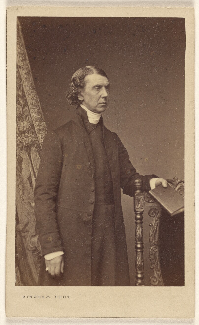 [Unidentified man standing, holding a book over a chair back]