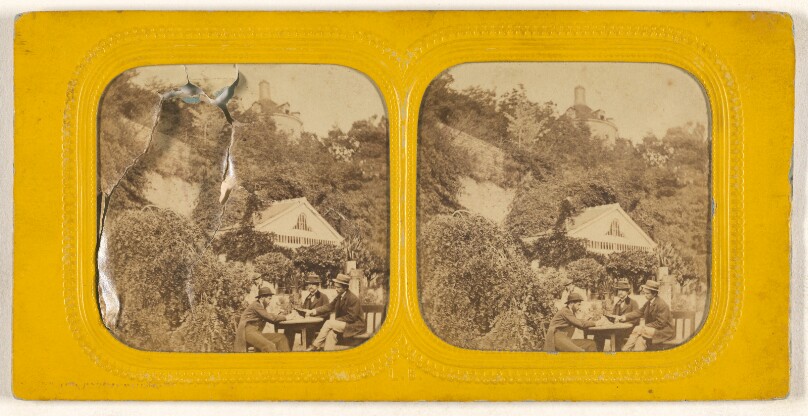 [Three men playing cards outside, houses and hillside in background]