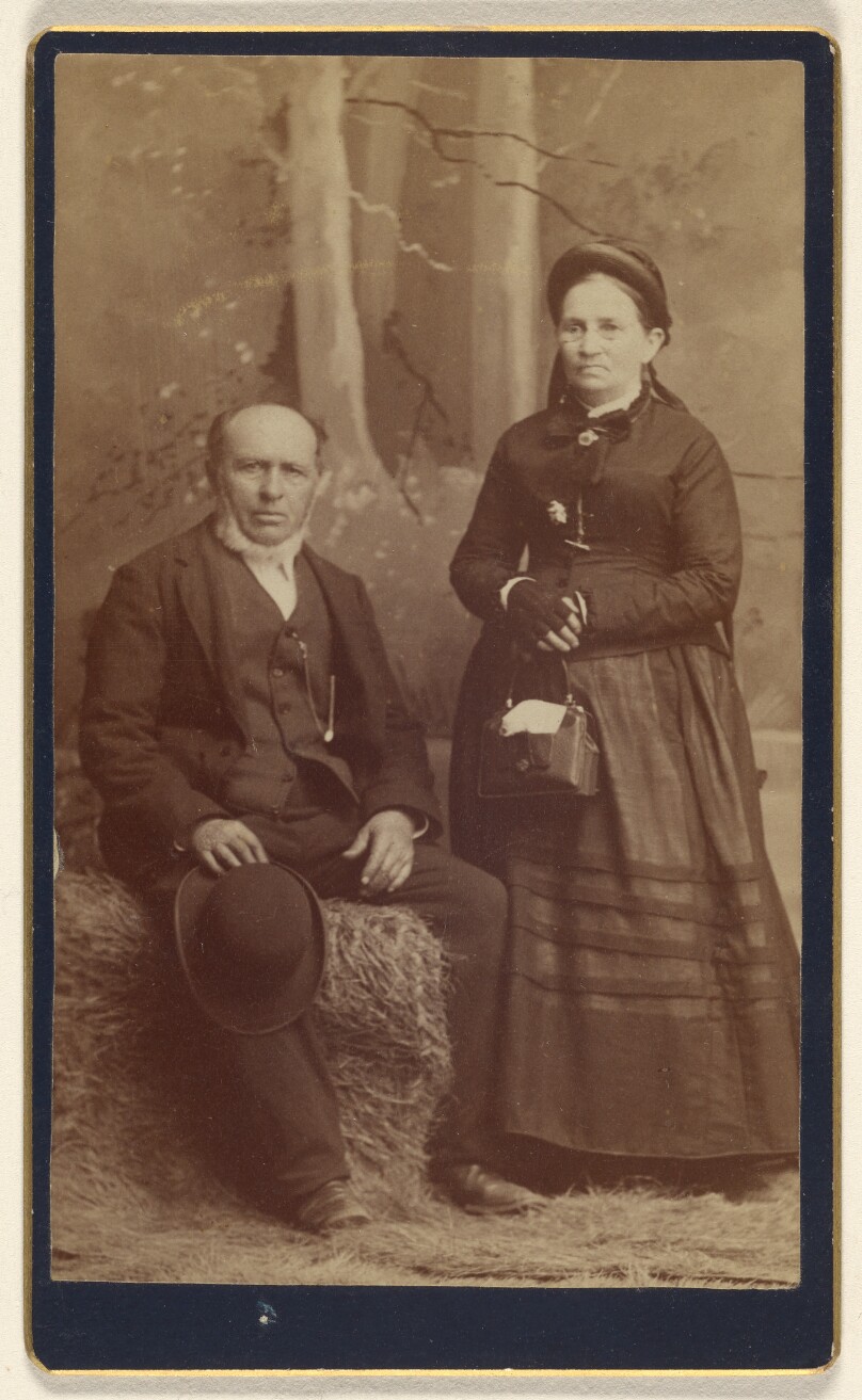 [Unidentified older couple: man seated in a bale of hay with a woman standing]