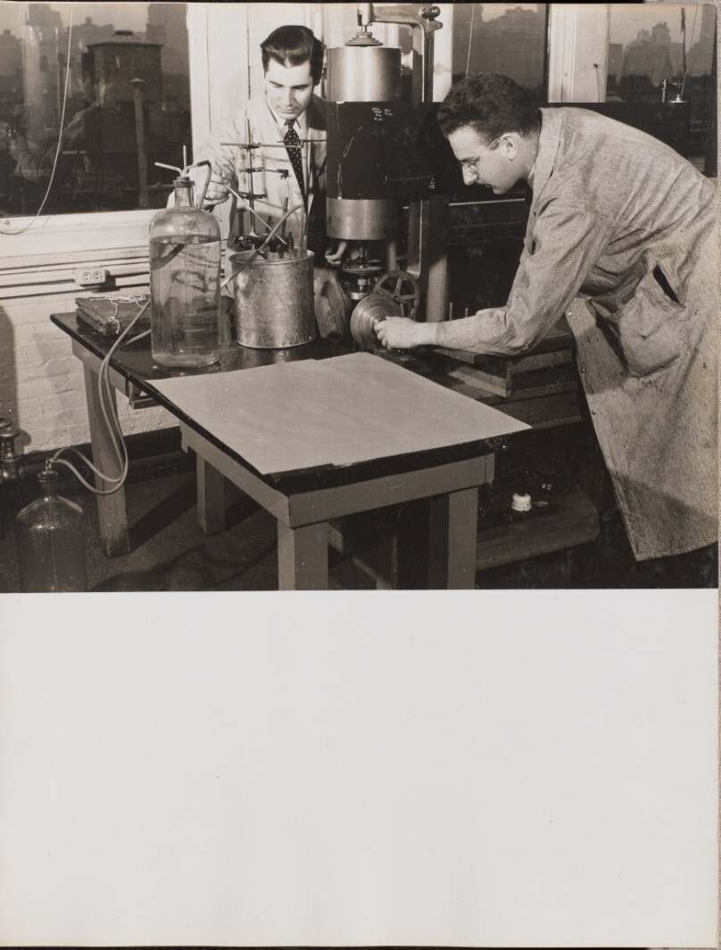 [Two men stand near a table with a large machine and a jug of liquid]