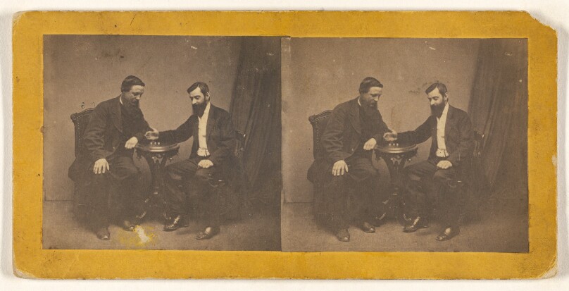 [Two men seated, posed at a table in a studio]