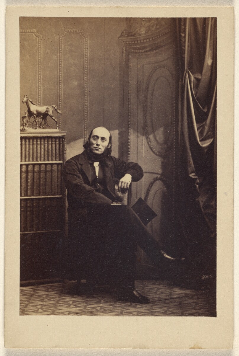 [Unidentified man seated with his legs crossed, holding a top hat]