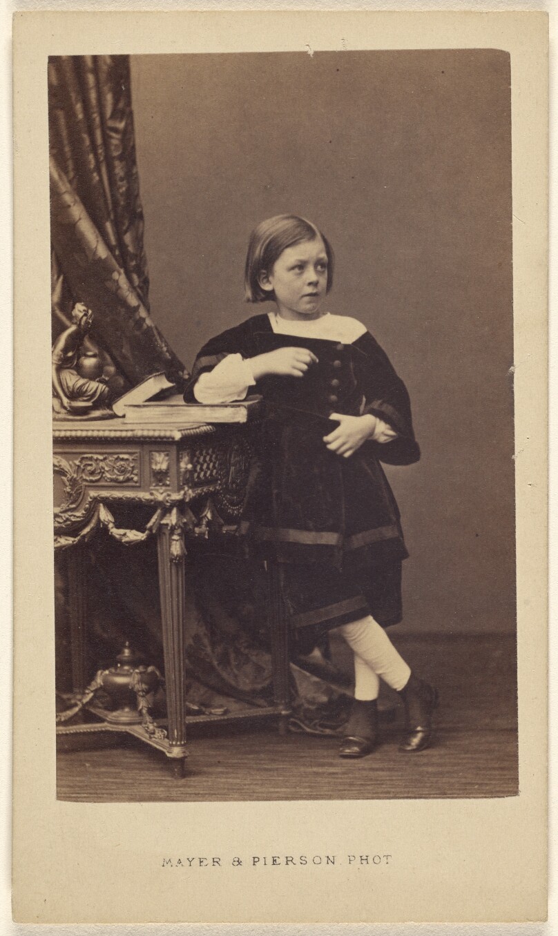 [Unidentified little girl standing, elbow on top of a closed book on a table]