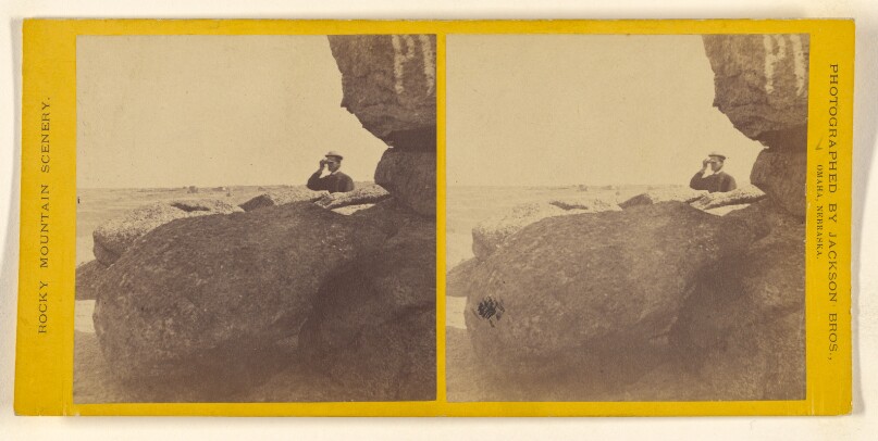 [Man wearing cap with hand to bill, looking out from the rocks]