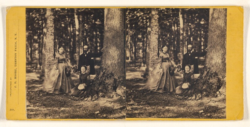 [Three unidentified people in a forest setting: woman and man standing, another woman seated near a tree]