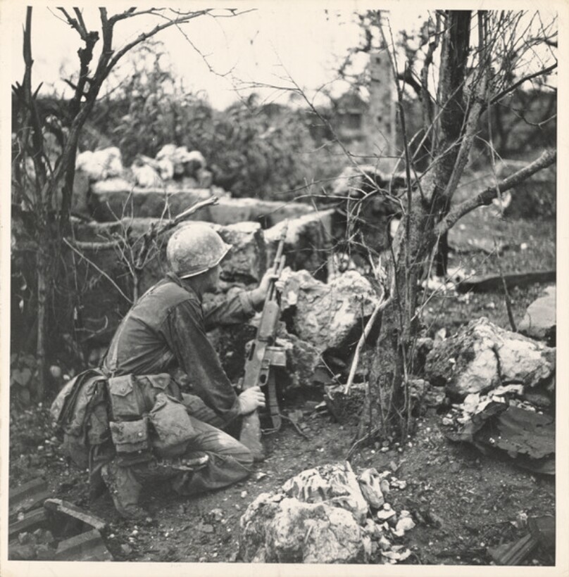 American infantryman Terry Moore holding his BAR rifle while crouching behind rocks as he assesses his attack path towards his objective, the coastal town of Yonaburu