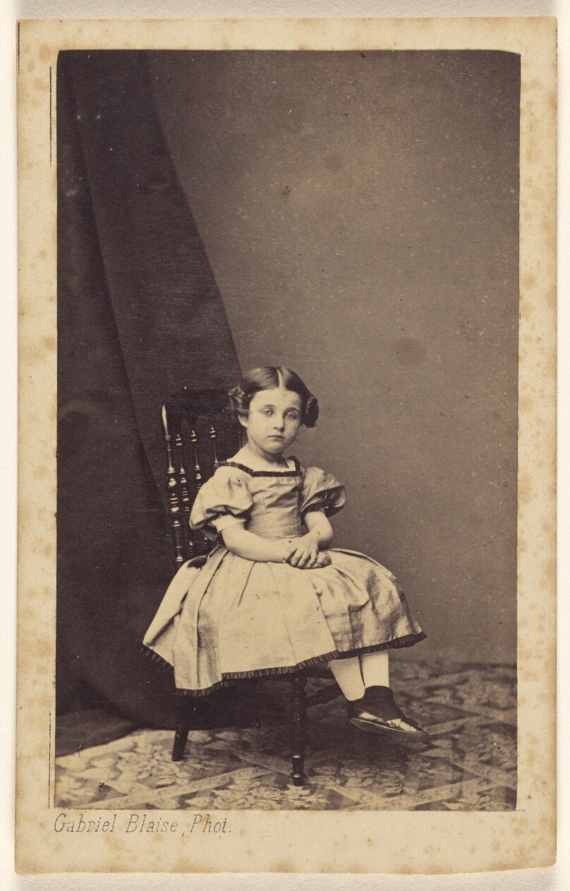 [Unidentified little girl with hair up on both sides, seated, with hands clasped]