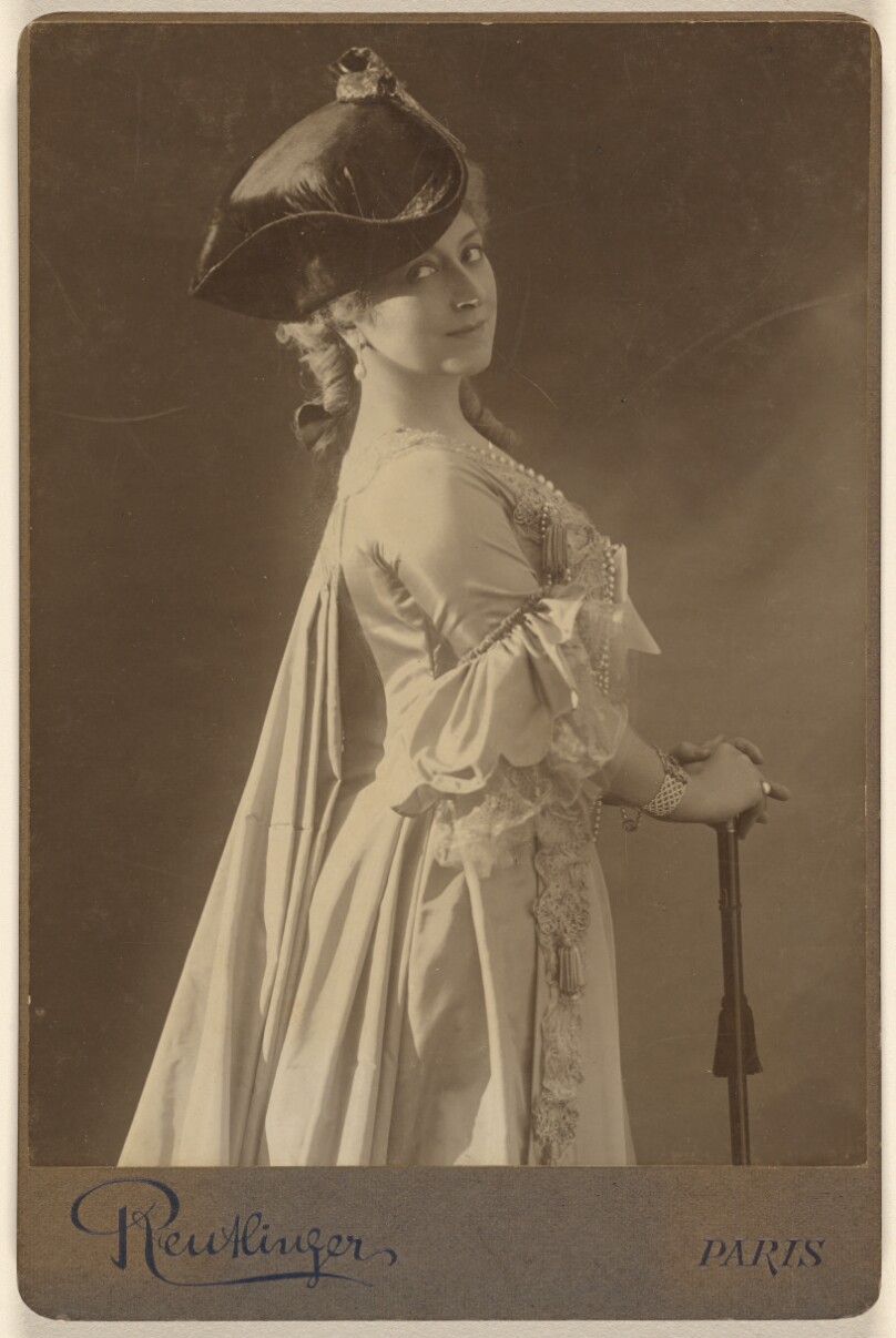 [Unidentified woman wearing a dark hat, standing, with hands resting on a walking stick]