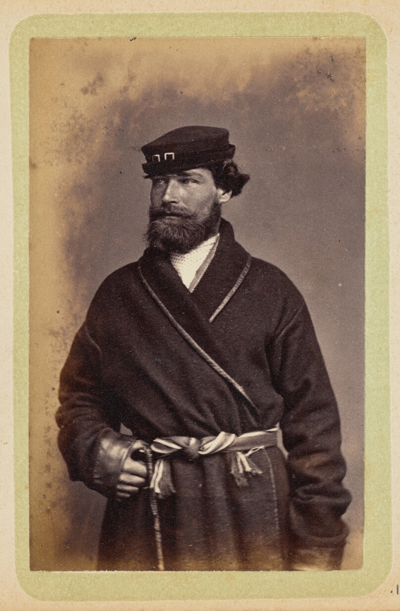 [Man posing in dark coat and sash]