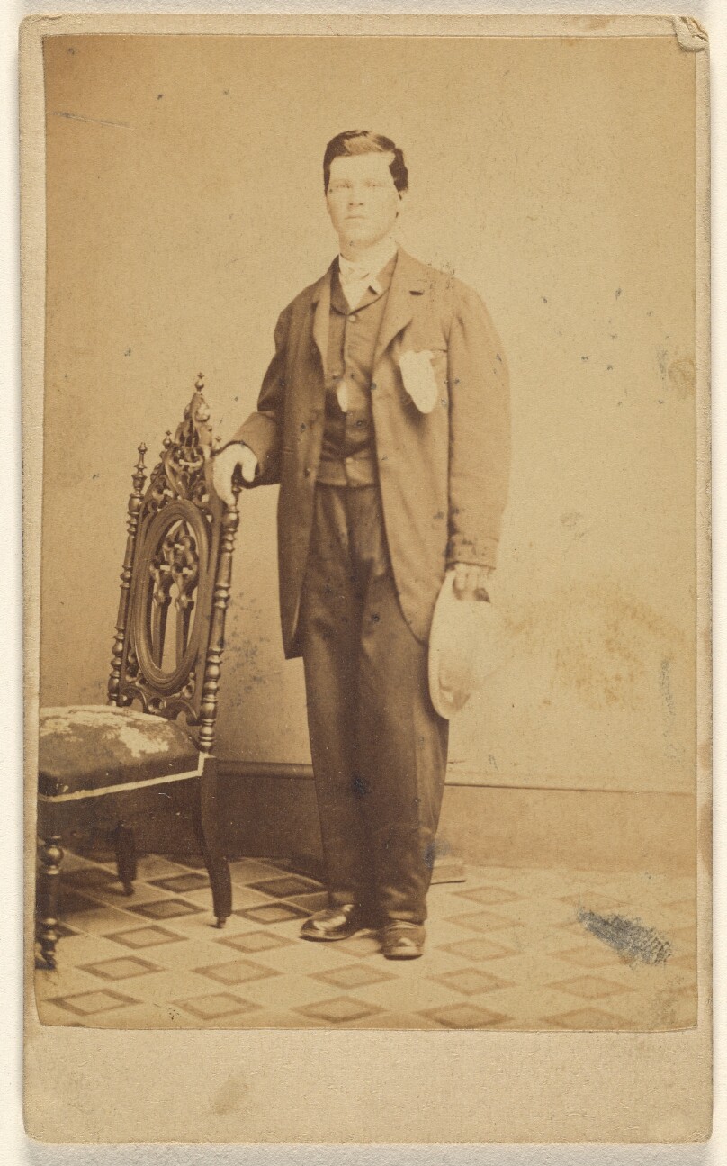 [Unidentified man standing holding his hat in one hand and holding a chair back with the other]