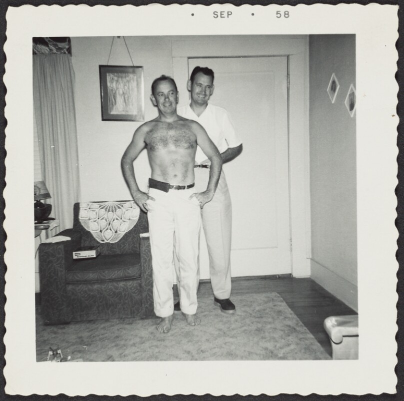 [Two men, one shirtless, in a living room]