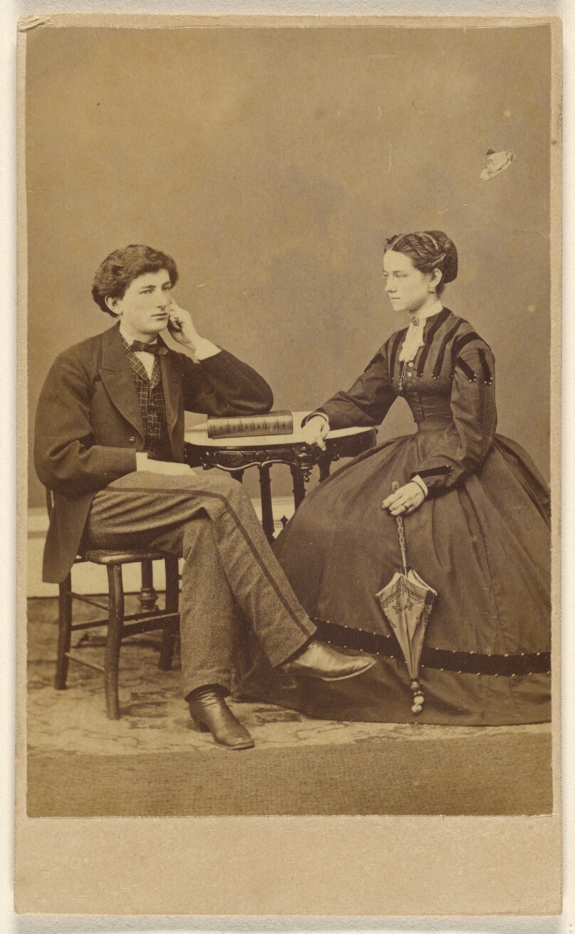 [Unidentified couple seated: man with hand to cheek, elbow on book; woman holding a parasol]
