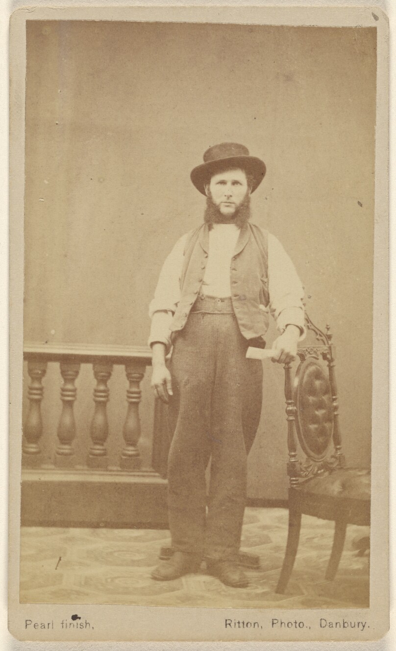 [Unidentified man with long, bushy muttonchops wearing a hat, standing, with one arm on a chair back]