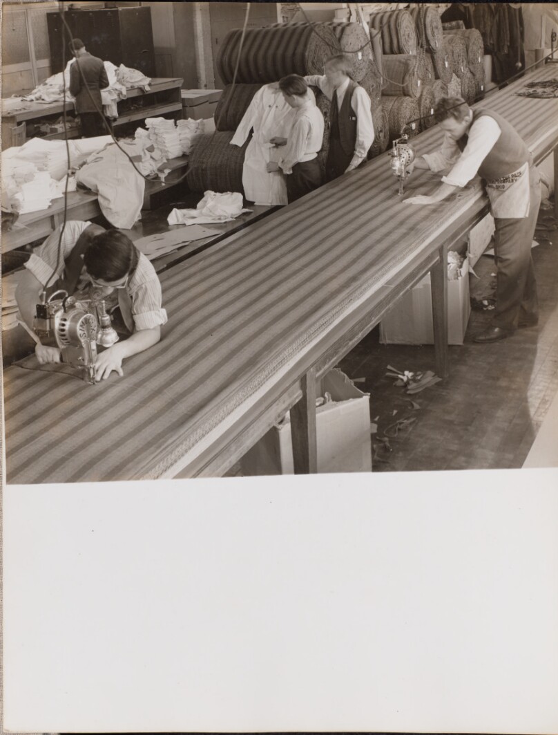 [Two men wearing aprons cut fabric]