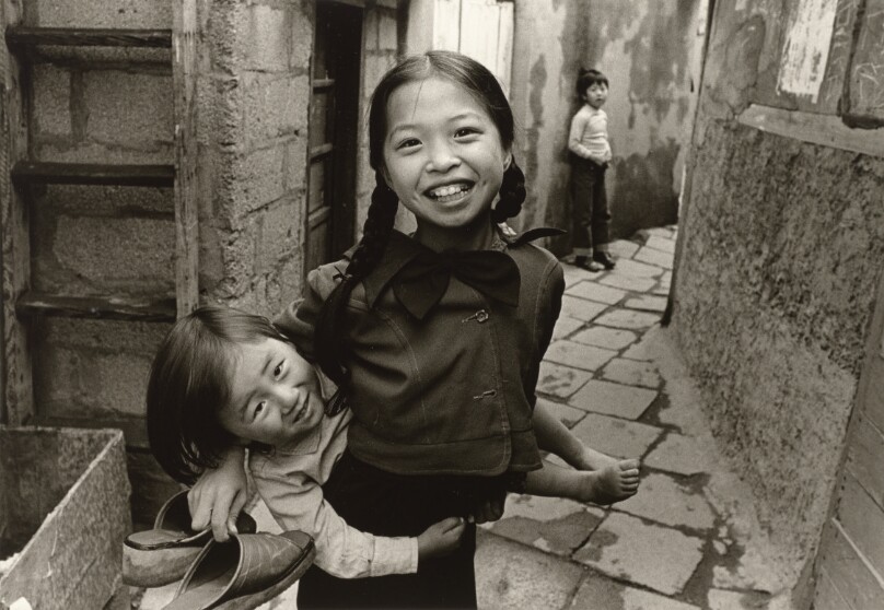 [Smiling Girl Carrying a Smaller Girl Sideways on Her Back, Young Boy Standing in the Alley]