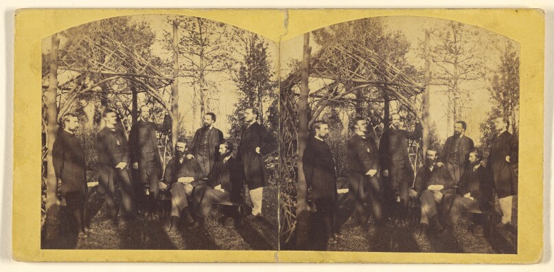 [Group of bearded men outside among the pine trees and open-thatched gazebo]