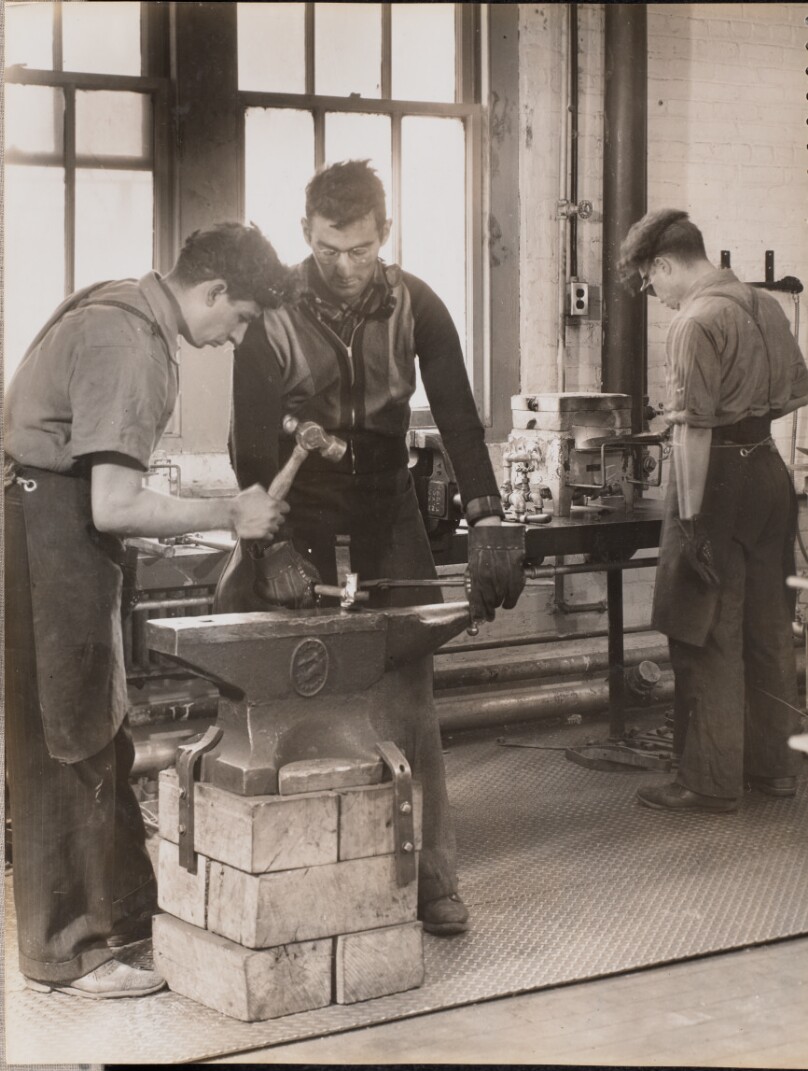 [Two men hammer metal on an anvil]
