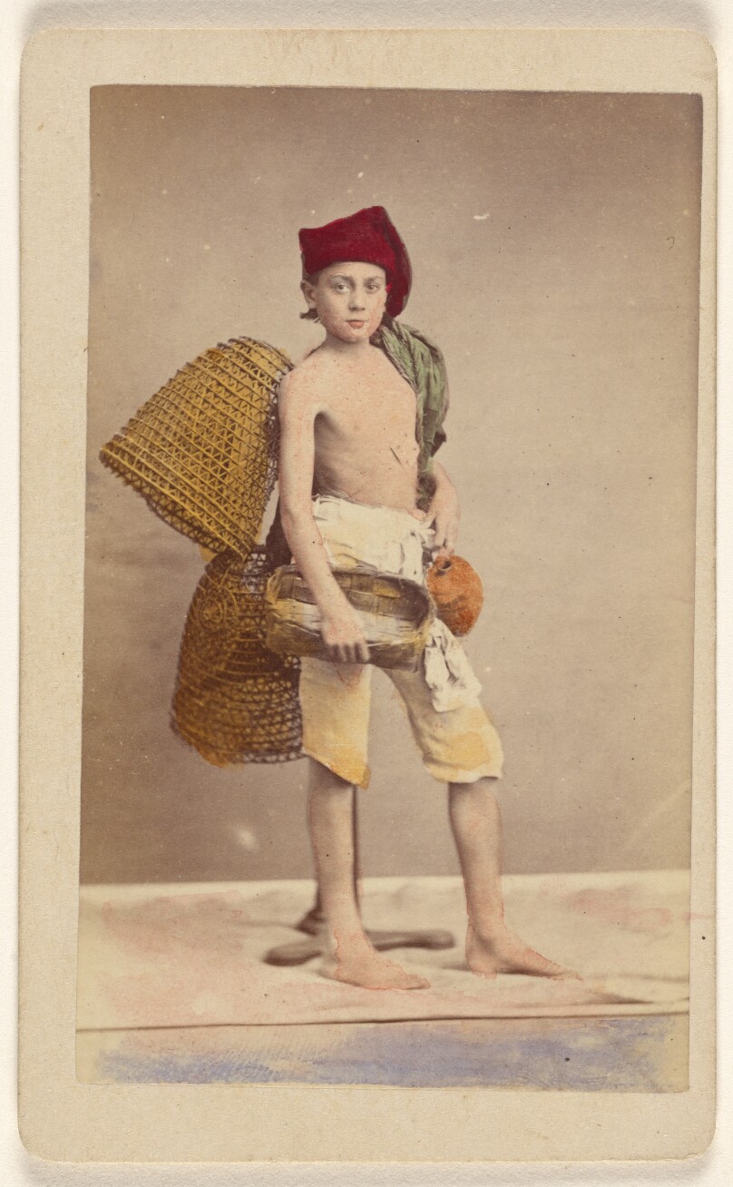 [Unidentified boy wearing native costume with baskets on back and carrying a water jug and small basket, standing]