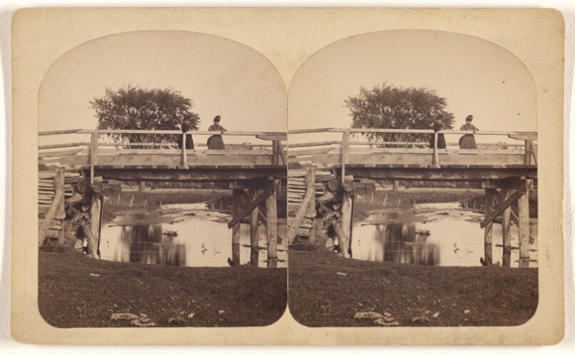 [Two women standing on a wooden bridge, shallow stream underneath]