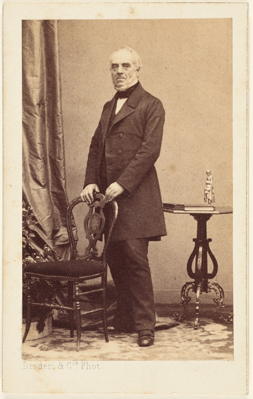 [Unidentified elderly man standing, holding the back of a chair]