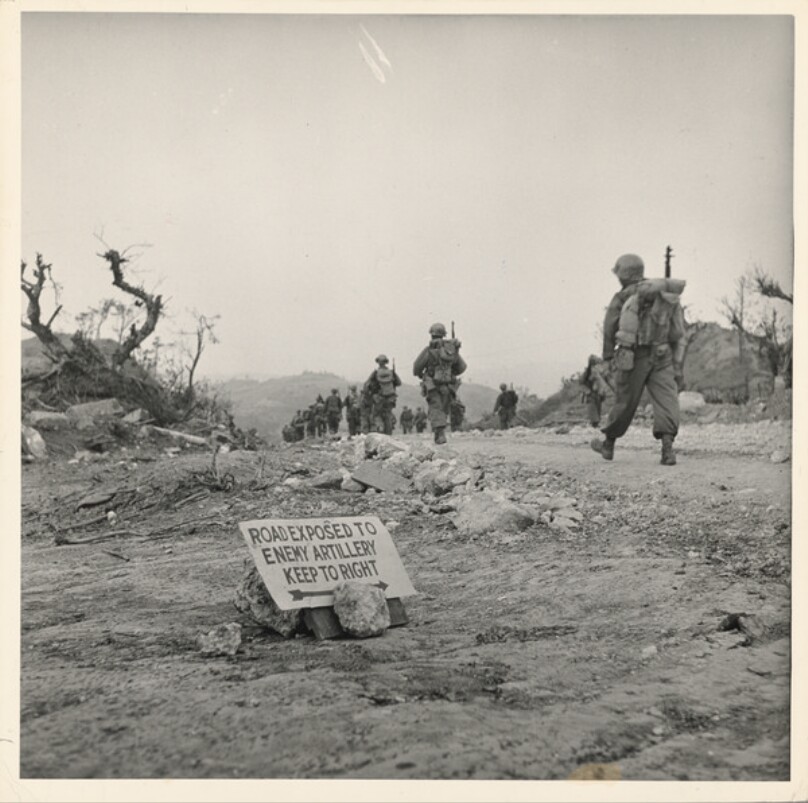 US Marines march past sign reading ROAD EXPOSED TO ENEMY ARTILLERY, KEEP TO RIGHT on their way towards the front lines during the fight to take Okinawa