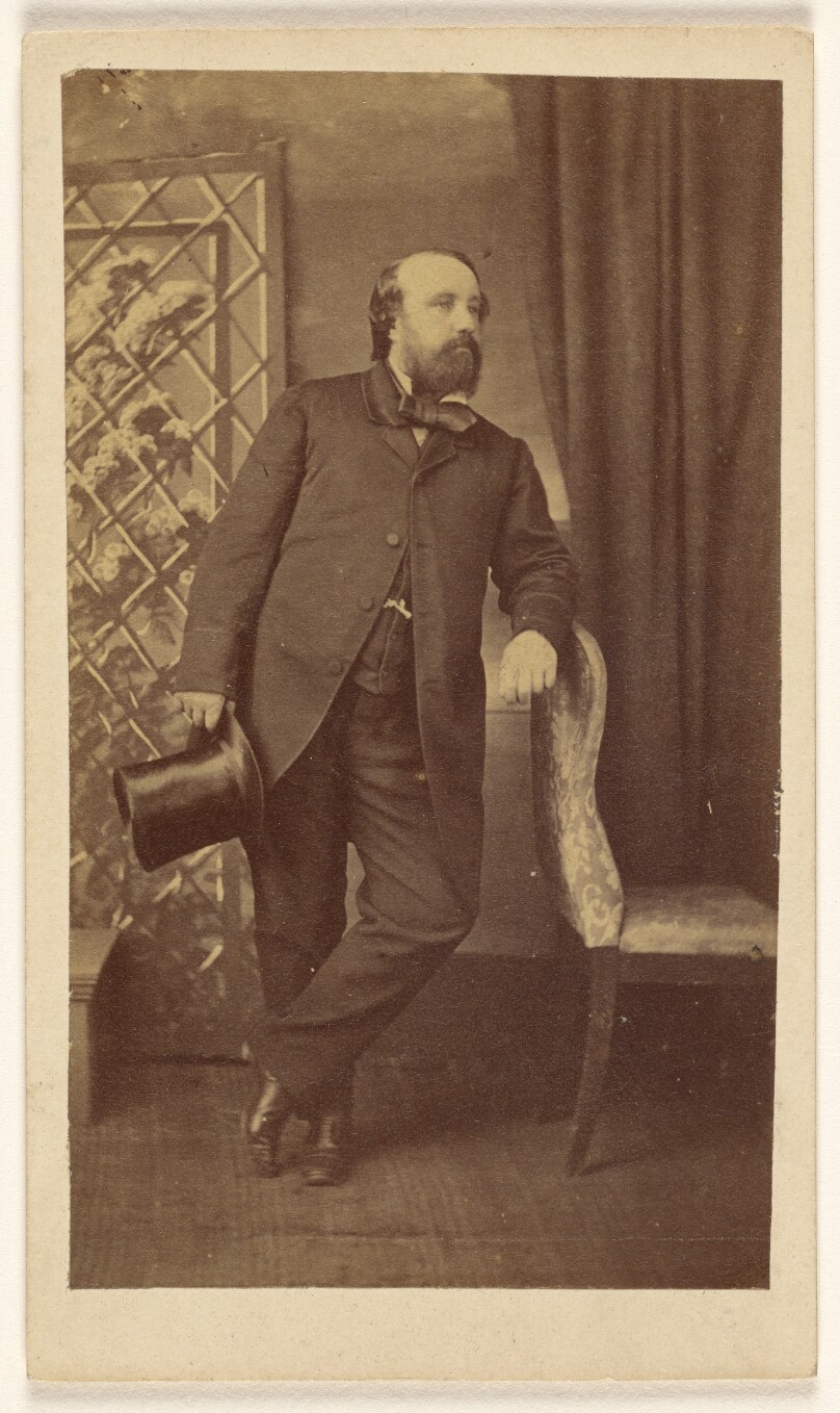 [Full-bearded man holding a top hat, standing, leaning on chair back]