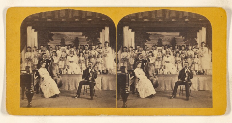 [Cast of a play on stage, two male musicians seated in front with well-dressed woman]