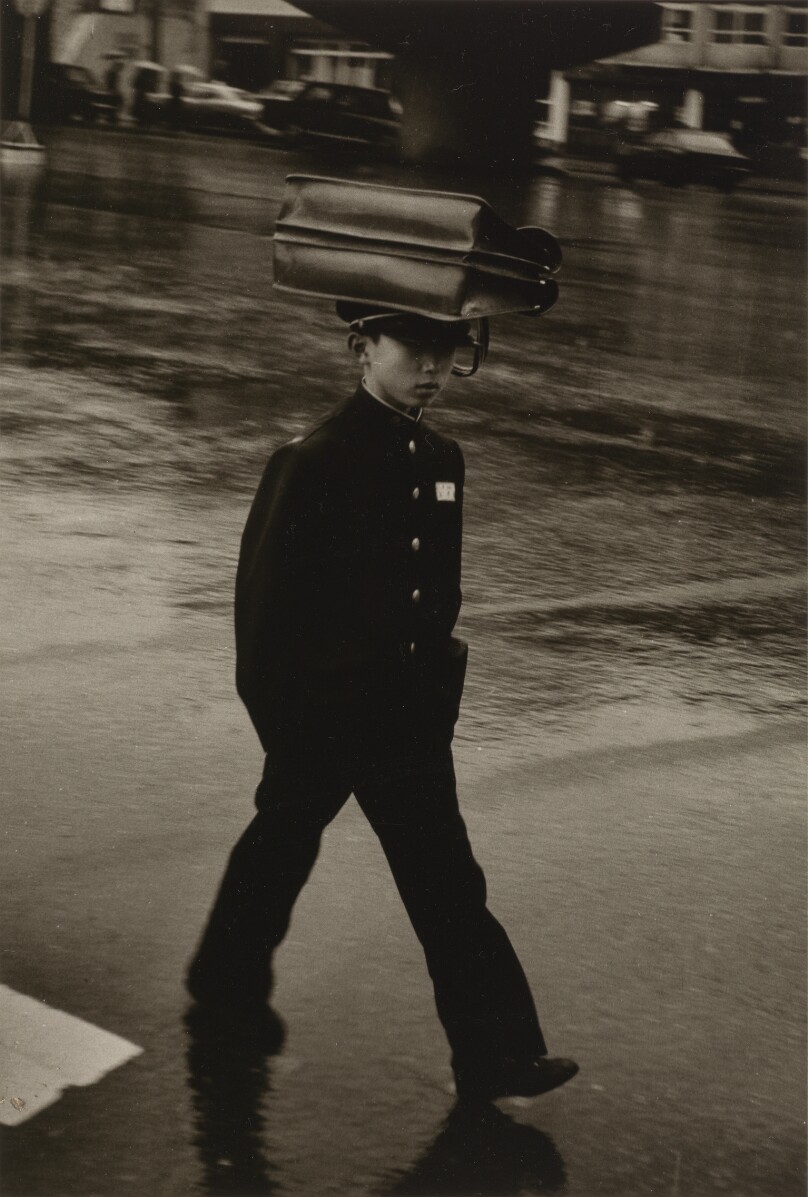 [School Boy in Uniform Walking Through the Rain with a Briefcase on Balanced on His Head]
