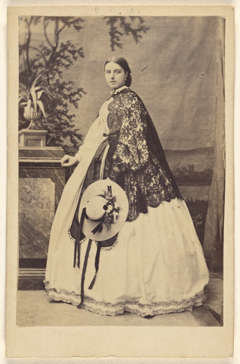 [Unidentified woman wearing a long dress with a dark shawl, standing, holding a bonnet]